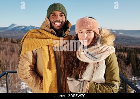 Joyeux jeunes dates interculturelles en vêtements d'hiver debout contre le ciel bleu au-dessus des montagnes couvertes de neige pendant le refroidissement le week-end d'hiver Banque D'Images