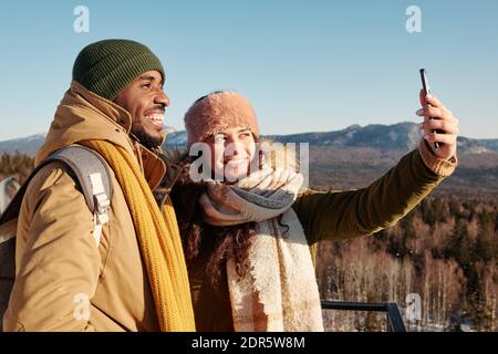 Joyeux jeune dates interculturelles dans les vêtements d'hiver faisant selfie devant de la caméra contre le ciel bleu au-dessus des montagnes couvertes de neige Banque D'Images