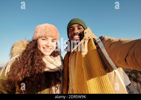 Joyeux jeune dates interculturelles dans les vêtements d'hiver faisant selfie devant de la caméra contre le ciel bleu le jour d'hiver ensoleillé pendant se détendre Banque D'Images