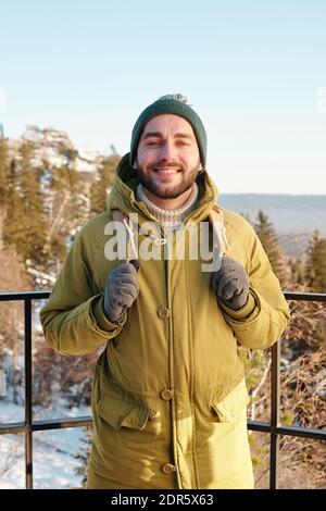 Jeune homme souriant avec sac à dos devant l'appareil photo contre les arbres et les rochers couverts de neige et de bleu clair ciel au-dessus le jour d'hiver Banque D'Images