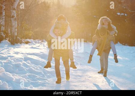 Deux types interculturels joyeux donnant à leurs amies pigegyback tout en déplaçant le week-end, le long de la route couverte de neige en forêt d'hiver Banque D'Images