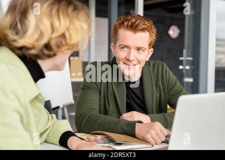 Jeune développeur souriant d'applications mobiles regardant une collègue pendant remue-méninges et discussion d'idées créatives lors de la réunion de démarrage Banque D'Images