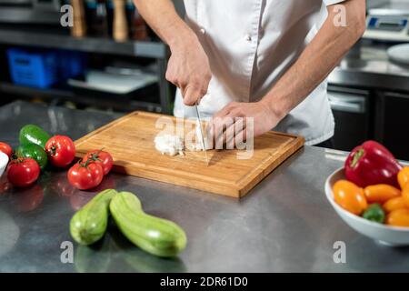 Mains de jeune chef masculin en uniforme coupant des oignons frais sur bois entouré de tomates et de courgettes pendant la cuisson déjeuner Banque D'Images