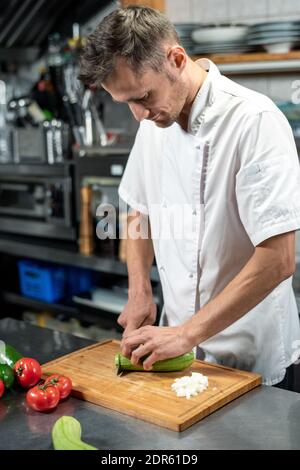 Jeune chef masculin en uniforme se pliant sur la table pendant la coupe courgettes vertes fraîches sur bois entouré de tomates et avocat Banque D'Images