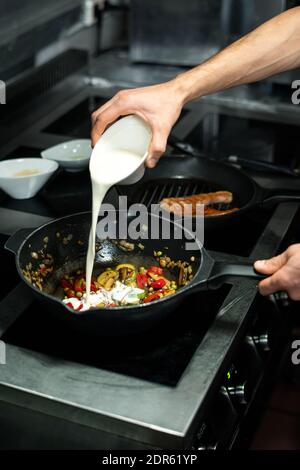 Main du chef cuisinier qui verse du lait frais dans une poêle avec un ragoût de légumes composé de pois verts, de poivrons, de champagnes, de courgettes et d'oignons Banque D'Images