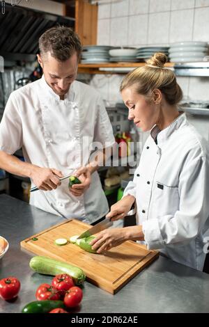 Jeune chef masculin professionnel avec couteau épluchant avocat pendant son femme stagiaire coupant des courgettes fraîches sur une planche en bois dans le cuisine Banque D'Images