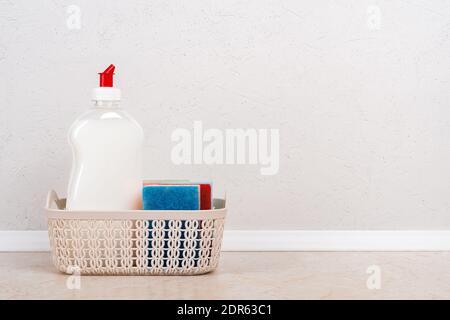 Liquide vaisselle blanc et peu d'éponges mousse dans un petit panier sur la table de cuisine contre un mur en béton blanc. Détergent ménager. Copier l'espace. Banque D'Images