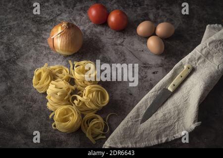 Un gros plan d'oignons frais, d'œufs, de tomates et de pâtes crues avec couteau et nappe sur une table en pierre Banque D'Images