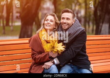 Charmant embrassant heureux couple assis sur le banc romantique embrassé dans le parc portant des manteaux et des foulards collectant un bouquet de feuilles mortes. Amour Banque D'Images