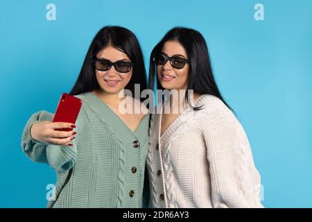 Des jumeaux en lunettes 3D, dans les mêmes vêtements, prennent un selfie sur un smartphone rouge. Arrière-plan bleu. Banque D'Images