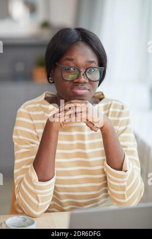 Portrait vertical de la jeune femme afro-américaine portant des lunettes et regardant à la caméra tout en étant assis au bureau dans un intérieur confortable Banque D'Images