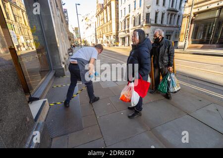 Un homme applique du ruban rayé au sol à l'extérieur d'un magasin de jetons dans le centre de Manchester comme guide social de distanciation. Les clients portant un EPI attendent Banque D'Images