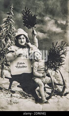 Mère et petit garçon avec traîneau, cadeaux, arbre de Noël. Carte postale allemande vintage avec grain de film original et rayures Banque D'Images