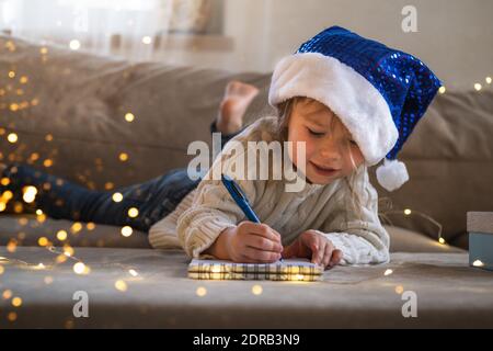 Jeune fille en chapeau de père Noël bleu écrivant une lettre de souhait au Père Noël. Concept de Noël ou de nouvel an. Banque D'Images