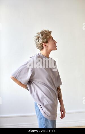 Vue latérale de la femme androgyne dans le t-shirt et jean tenant la main à la taille et regardant loin contre le blanc mur à la maison Banque D'Images