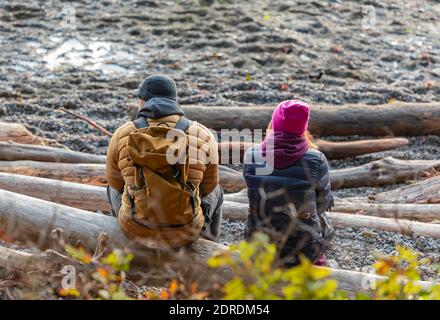 Un jeune couple assis sur la rive au bord de la mer, appréciant le coucher du soleil lors d'une soirée hivernale froide à Vancouver Ouest. Photo de voyage, vue sur la rue, mise au point sélective. Banque D'Images