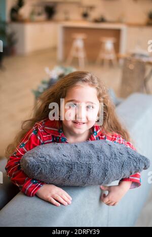 Jolie petite fille avec des yeux bleus et de longs cheveux en pyjama rouge se trouve sur le canapé gris dans la salle de séjour, embrasse un oreiller et sourit Banque D'Images