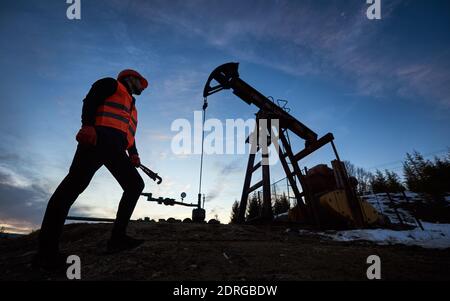 Faible angle de l'ouvrier pétrolier mâle se tenant près du cric de la pompe à pétrole sous le beau ciel de la soirée. Ingénieur pétrolier en travail gilet et casque tenant une clé industrielle et regardant la machine à bascule de pompe à huile. Banque D'Images