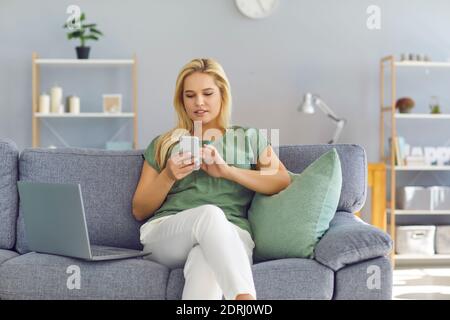 Jeune femme assise sur un canapé avec un ordinateur portable à la maison et utilisation de l'application téléphone mobile Banque D'Images