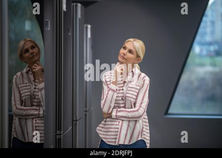 Femme dans une chemise rayée choisissant le réfrigérateur dans une salle d'exposition et il a l'air bien pensé Banque D'Images