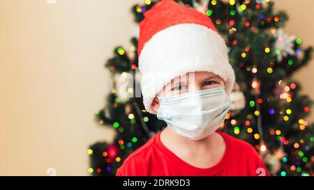 Garçon en chapeau rouge portant un masque médical. Arbre de Noël décoré sur fond. Copier l'espace Banque D'Images