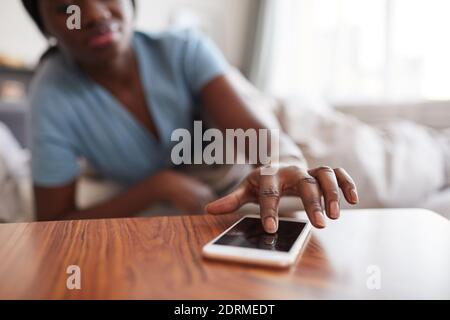 Gros plan d'une femme afro-américaine méconnue qui touche un smartphone sur une table de chevet tout en éteignant le réveil ou en vérifiant les messages le matin, co Banque D'Images