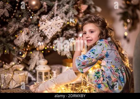 Une belle fille aux cheveux longs et bouclés est assise près d'un arbre de Noël. Tenant une guirlande de lumières de Noël. Le concept des loisirs d'hiver à hom Banque D'Images