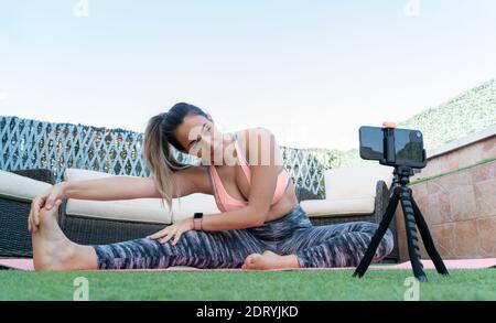 Jeune professeur de yoga enregistrant un cours en ligne avec son téléphone mobile dans le jardin de sa maison. Elle s'étire les jambes tout en regardant l'appareil photo Banque D'Images