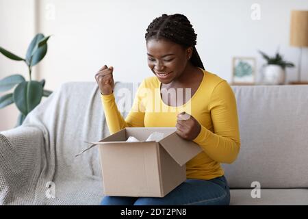 Une femme noire heureuse qui déboîte le colis à la maison réagit émotionnellement au succès des achats Banque D'Images