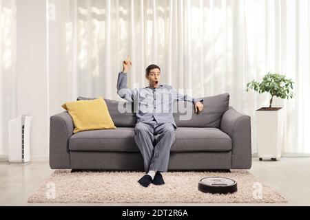 Jeune homme excité en pyjama assis sur un canapé à un aspirateur-robot sur le est orienté vers le haut sol Banque D'Images