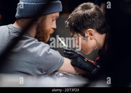 Un tatouage artiste avec des cheveux courts et des piercings se prépare à obtenir un tatouage sur le bras d'un homme dans un salon de tatouage. Banque D'Images