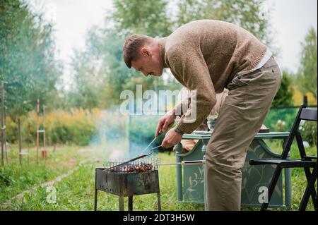 Le mari, le chef de famille, prépare des steaks et un barbecue pour sa femme lors d'un dîner à l'extérieur. Sur la pelouse dans la cour d'une maison de campagne Banque D'Images