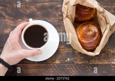 Petit-déjeuner le matin avec une tasse de café et un rouleau de cannelle sur fond de bois. La main d'un homme tient le café. Banque D'Images