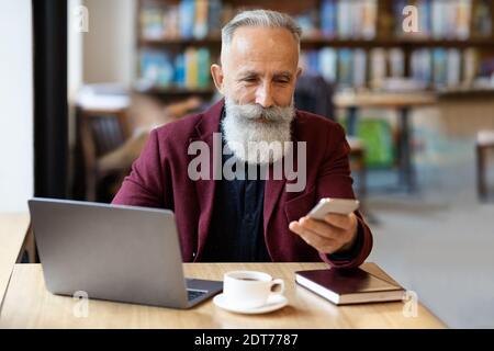 Un riche entrepreneur âgé avec un smartphone assis devant un ordinateur portable Banque D'Images