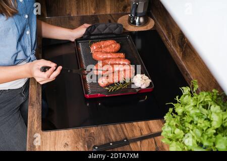 Femme cuisant des saucisses pour le déjeuner sur la cuisine, vue rapprochée.la fille prépare des saucisses sur une poêle à rôtir sur une cuisinière. Banque D'Images