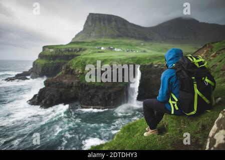 Danemark, Iles Féroé, Gasadalur Village, MÂ·Lafossur Waterfall, Homme avec sac à dos assis au bord de la falaise et regardant la cascade de Mulafossur Banque D'Images