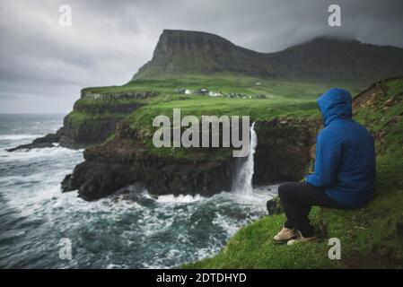 Danemark, Îles Féroé, Gasadalur Village, MÂ·Lafossur Waterfall, Homme assis au bord de la falaise et regardant la cascade de Mulafossur Banque D'Images