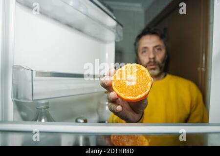 Homme d'âge moyen présentant la moitié de l'orange frais à l'appareil photo en vous tenant près de la porte ouverte du réfrigérateur vide Banque D'Images