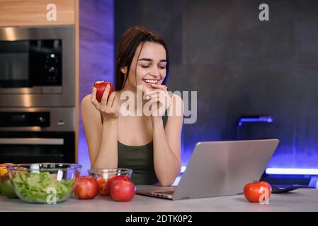 Belle jeune femme mange de la pomme rouge assis dans une cuisine moderne et utilise un ordinateur portable. Jolie fille regarde un blog intéressant d'Internet. Banque D'Images