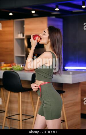 Jolie jeune femme savourant une pomme dans la cuisine après un délicieux petit déjeuner sain. Mode de vie sain et concept végétarien. Banque D'Images