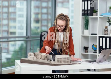 Architecte féminin expérimenté et élégant avec des cadenas travaillant avec un modèle en carton de zone résidentielle dans le bureau architectural Banque D'Images