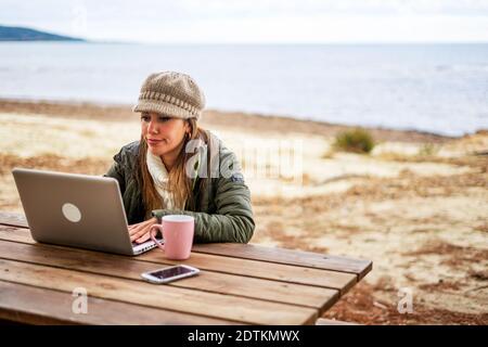 Internet femme freelance travaillant à l'extérieur dans la station balnéaire assis à table en bois portant des vêtements automne-hiver et chapeau de laine avec thé tasse près de son alu Banque D'Images