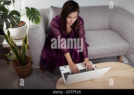 Travailler à domicile. Bureau à domicile. Femme d'affaires dans une chemise de nuit en satin violet et peignoirs de dentelle en utilisant l'ordinateur portable au milieu de la nuit. Banque D'Images