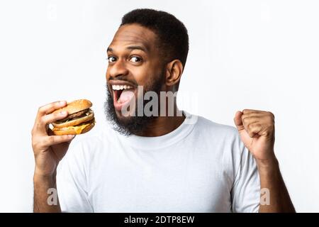 Joyeux Black Guy bite Burger appréciant un repas chaud, fond blanc Banque D'Images