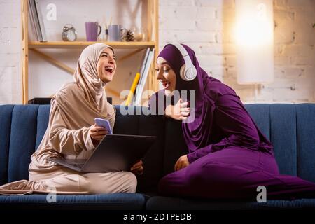 Rire. Heureux et jeunes deux musulmanes à la maison parlant, souriant, s'amusant, assis sur un canapé. Amitié, technologie moderne, émotions concept. Style de vie et culture, traditions arabes, ethnie. Banque D'Images