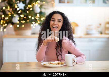 Collation de Noël. Femme joyeuse appréciant des biscuits de vacances et du chocolat chaud dans la cuisine Banque D'Images