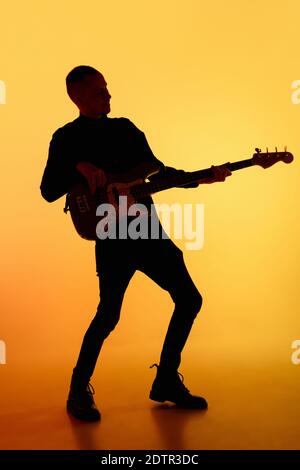 Fête. Silhouette de jeune guitariste mâle isolée sur fond de studio de gradient orange à la lumière du néon. Des ombres magnifiques en action, en action. Concept des émotions humaines, de l'expression, de la publicité, de la musique, de l'art. Banque D'Images