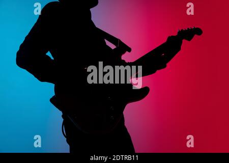 Lumineux. Silhouette de jeune guitariste mâle isolée sur fond de studio dégradé bleu-rose au néon. Des ombres magnifiques en action, en action. Concept des émotions humaines, de l'expression, de la publicité, de la musique, de l'art. Banque D'Images