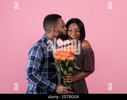 Un gars afro-américain affectueux embrassant sa copine heureuse et donnant son bouquet de roses sur fond rose studio Banque D'Images