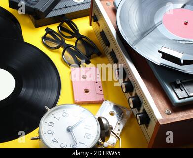 Attributs vintage, médias des années 80. Lecteur de vinyle, cassettes vidéo, cassettes audio, disques, radio, réveil rétro sur fond jaune. Banque D'Images